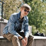 Fashionable young man in sunglasses and a denim jacket relaxing outdoors on a sunny day.